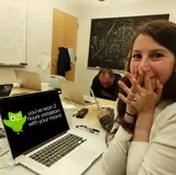 Woman covering her mouth in delight beside laptops and a chalkboard, laptop screen reading "you've won 2 hours visitation with your mom!"