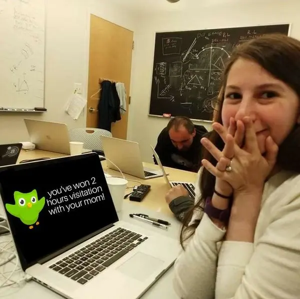 Woman covering her mouth in delight beside laptops and a chalkboard, laptop screen reading "you've won 2 hours visitation with your mom!"