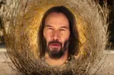 Film still of a bearded, long-haired man's face embedded in a large tumbleweed in a sunlit desert