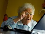 Elderly white-haired woman in a blue patterned blouse lifting her glasses to squint at an open laptop screen