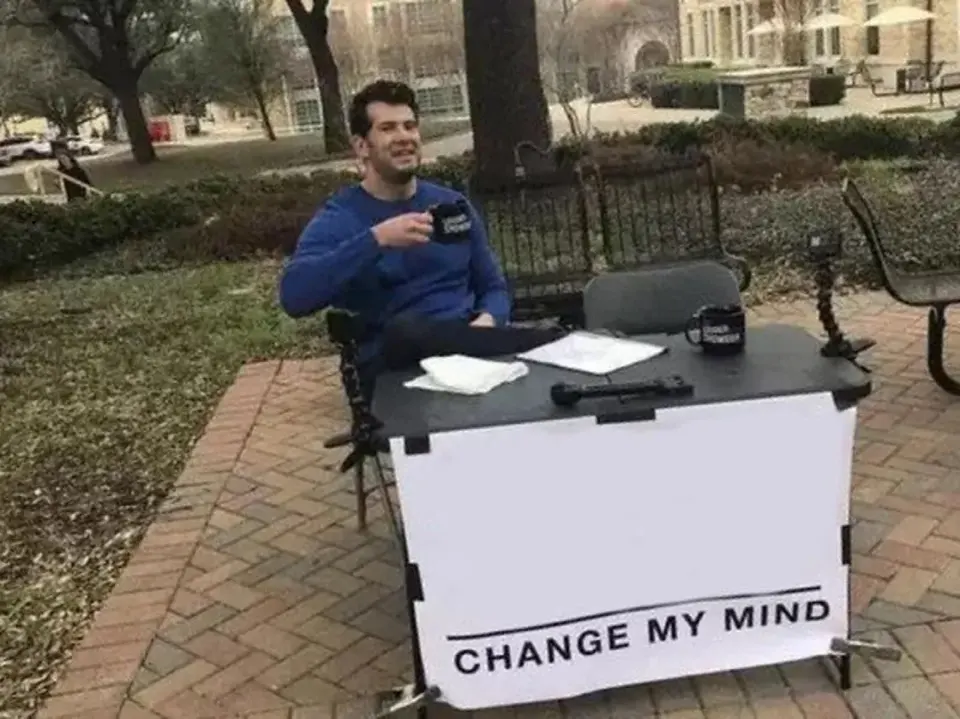 Man in a blue sweater seated at a folding table on a campus lawn behind a sign reading "CHANGE MY MIND"
