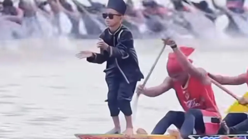 Young boy in black traditional dress, sunglasses and cap dancing on the prow of a racing boat while rowers paddle behind