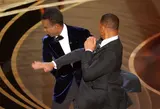 Two men in tuxedos on a lit awards stage, one swinging an open-handed slap into the other's face