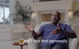 Bald man in a blue T-shirt gesturing during a living-room interview, whiskey and cigar nearby, captioned "...and I took that personally"