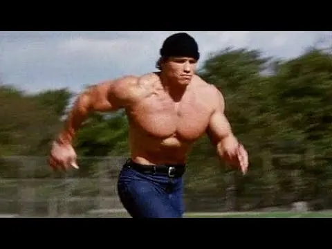 Film still of a shirtless, heavily muscled man in a dark beanie and blue jeans sprinting across a grassy field