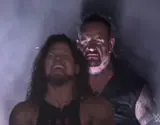 Two wrestlers in fog: a long-haired man grinning in front, a grim pale-faced man looming behind him, WWE logo