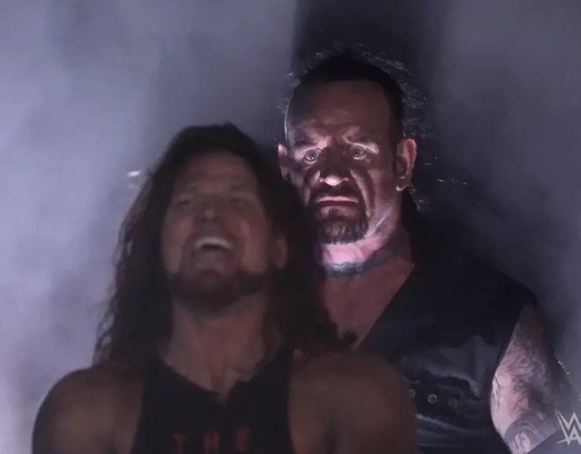 Two wrestlers in fog: a long-haired man grinning in front, a grim pale-faced man looming behind him, WWE logo