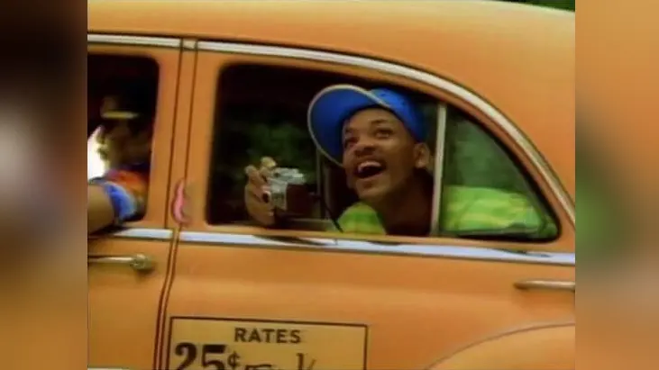 Grinning young man in a blue cap leaning out of an orange taxi window holding a camera, "RATES 25¢" sign
