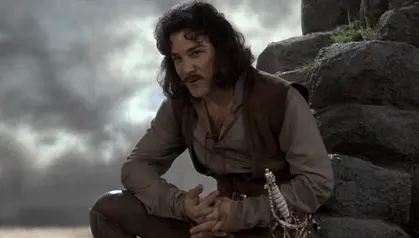 Long-haired mustachioed swordsman in a tan shirt crouching among grey rocks under a stormy sky, a sword hilt at his hip