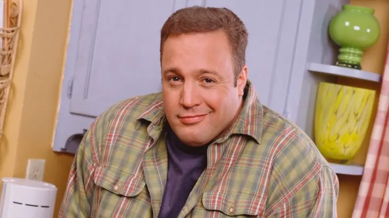 Heavyset man in a plaid shirt over a purple tee leaning forward with raised shoulders and a tight-lipped smirk